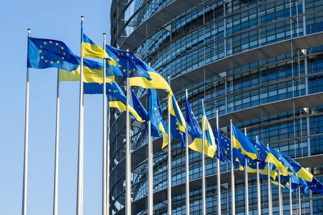 Mai multe steaguri ale Uniunii Europene și ale Ucrainei sunt arborate pe catargi înalți în fața unei clădiri moderne cu fațadă de sticlă sub un cer senin.