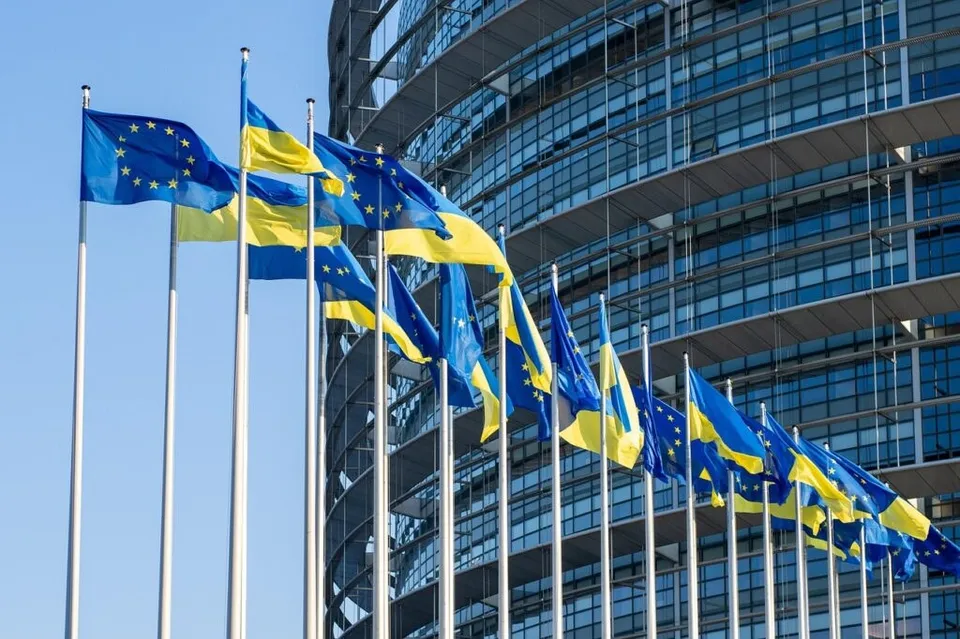 Mai multe steaguri ale Uniunii Europene și ale Ucrainei sunt arborate pe catargi înalți în fața unei clădiri moderne cu fațadă de sticlă sub un cer senin.