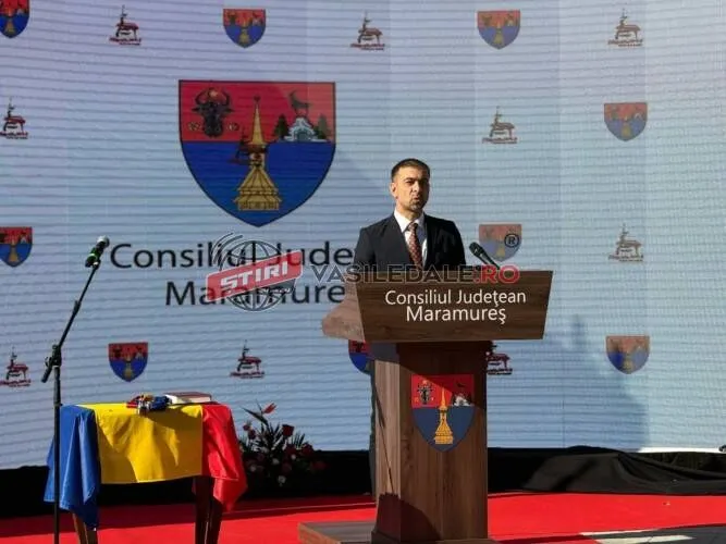 Un bărbat într-un costum închis la culoare stă în spatele unui podium de lemn pe o scenă cu un fundal alb decorat cu blazoane.