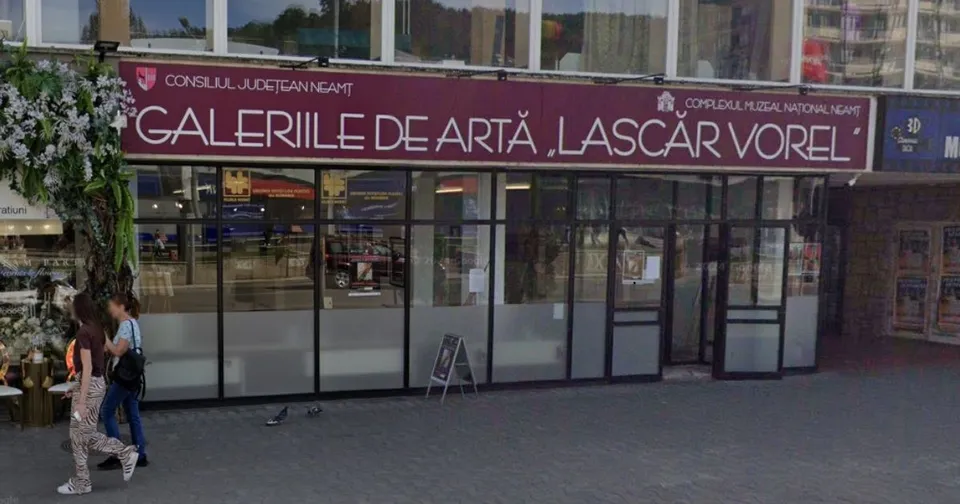 O persoană merge pe trotuar în fața unei clădiri cu vitrine mari pe care este afișat un panou cu textul Galeriile de Artă Lascăr Vorel.