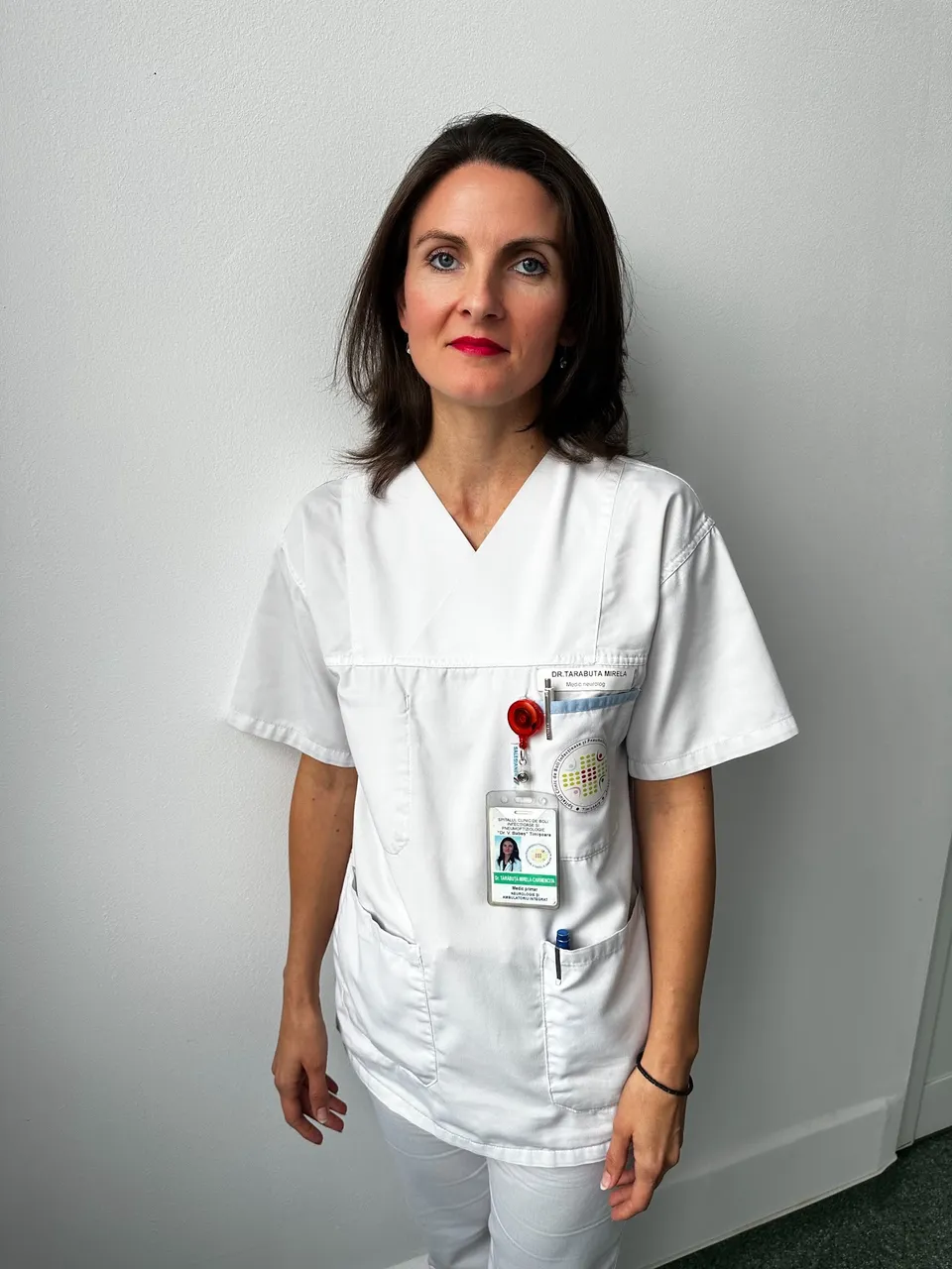 O femeie cu părul scurt și negru poartă o uniformă medicală albă cu un badge și un instrument medical atașat pe buzunarul de sus.