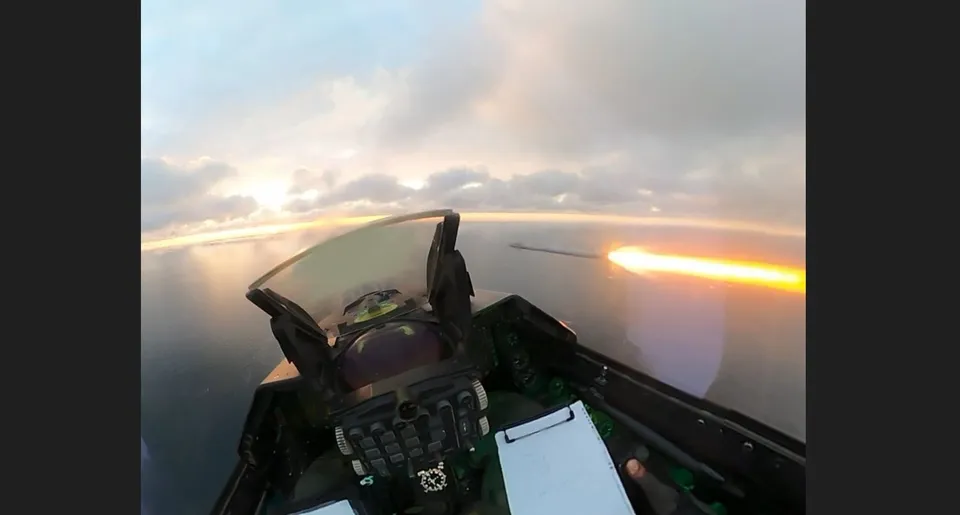 Perspectiva din interiorul unui avion de voadă arată un câmp de nori sub un cer luminos și o traiectorie luminoasă de foc în fundal.