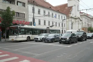 Un autobuz alb și gri circulă pe un drum asfaltat alături de mai multe mașini de culoare neagră și albă într-un oraș.