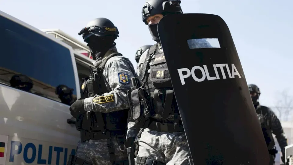 Patru polițiști în echipament tactice și casci, unul dintre ei ținând un scut negru pe care scrie POLITIA, stau lângă un vehicul alb.