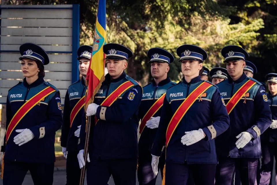 Un grup de polițiști în uniformă albastră cu bandiere colorate pe piept și căciuli albe stă în linie în fața unor copaci.