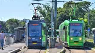 Două tramvaie moderne, unul gri și unul verde, sunt oprite pe șine lângă o stație de pasageri sub un cer senin.