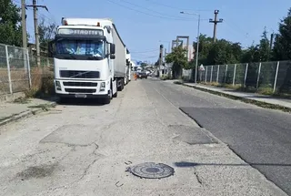 Un camion alb este parcat pe o stradă cu asfalt deteriorat, lângă un gard de sârmă și stâlpi de utilități sub un cer senin.