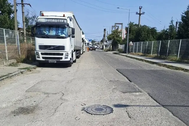 Un camion alb este parcat pe o stradă cu asfalt deteriorat, lângă un gard de sârmă și stâlpi de utilități sub un cer senin.