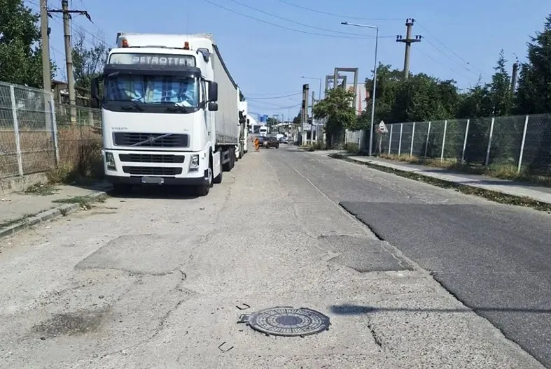Un camion alb este parcat pe o stradă cu asfalt deteriorat, lângă un gard de sârmă și stâlpi de utilități sub un cer senin.