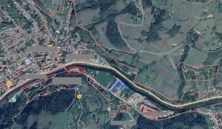 O imagine aeriană de satelit prezintă o zonă cu case, drumuri și terenuri verzi delimitată de linii roșii pe un fundal de pădure și dealuri.
