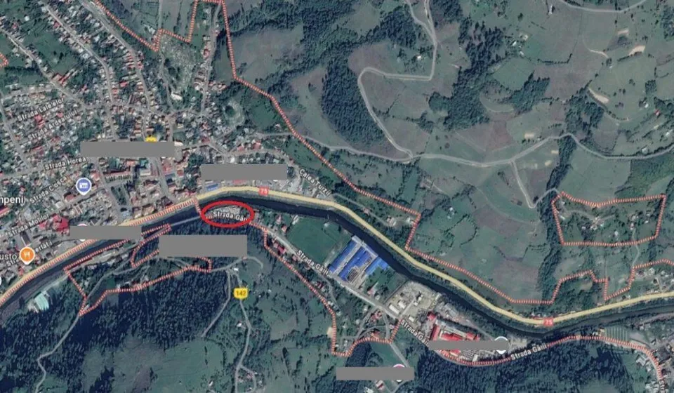 O imagine aeriană de satelit prezintă o zonă cu case, drumuri și terenuri verzi delimitată de linii roșii pe un fundal de pădure și dealuri.