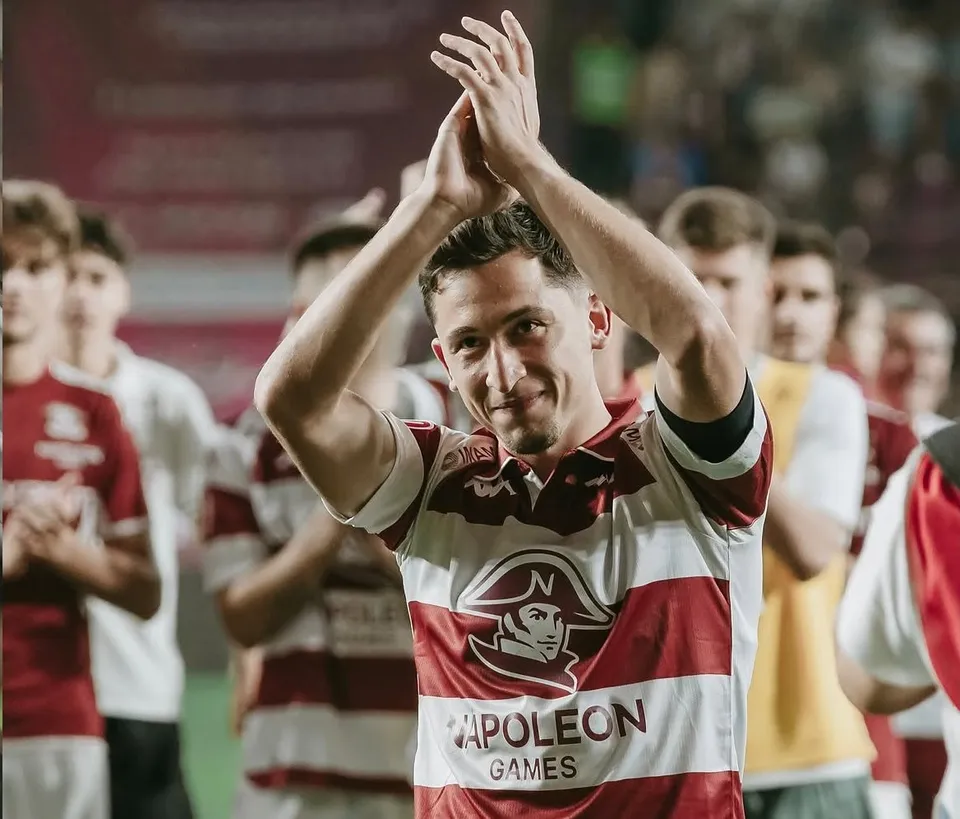 Un bărbat cu tricou alb și roșu clapă cu mâinile deasupra capului, fiind înconjurat de alți jucători pe un teren de fotbal.