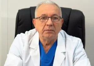 Un bărbat cu ochelari și părul cărunt poartă o halat albă peste un tricou albastru, stând pe un scaun de birou negru.