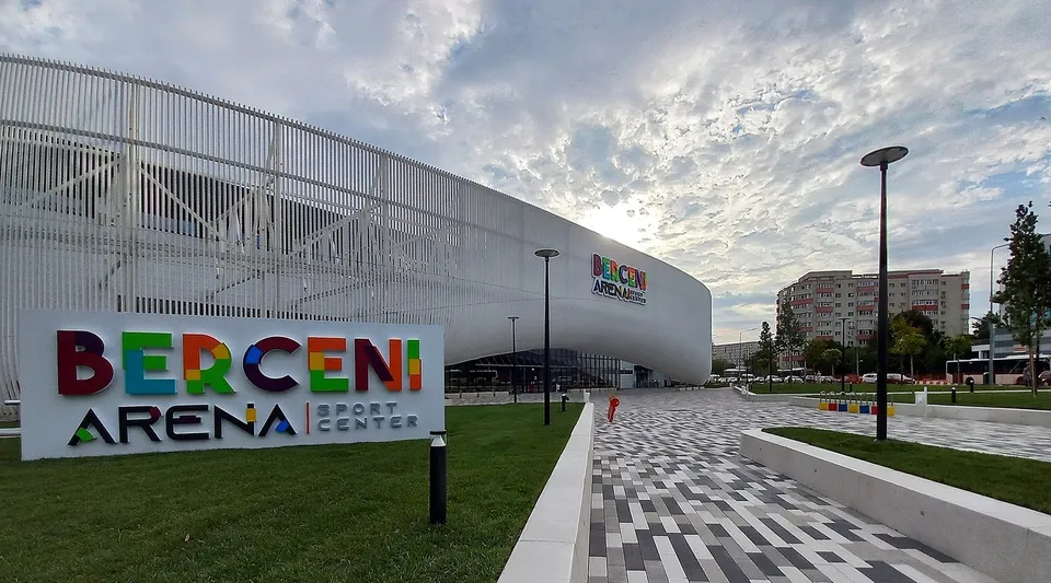 O clădire modernă cu fațadă gri și un panou publicitar colorat cu textul Berceni Arena Sport Center sunt situate lângă un drum pavat.