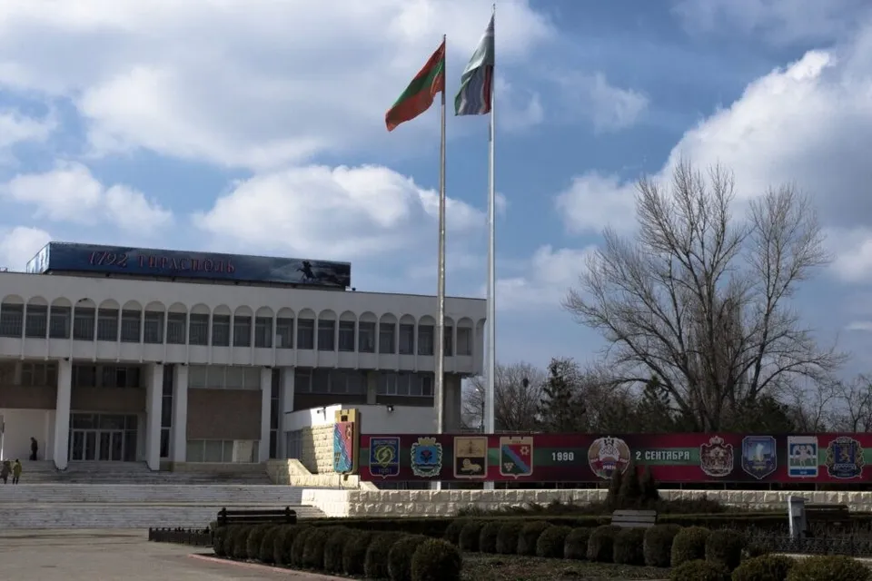 Două steaguri sunt ridicate pe două catargi înalte în fața unei clădiri albe sub un cer cu nori.