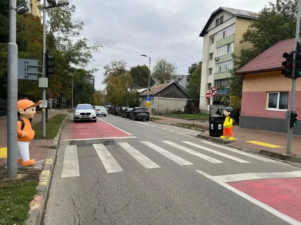 Un manechin de copil cu ghiozdan portocaliu stă pe marginea trotuarului lângă un semafor, lângă o trecere de pietoni marcată pe drum.