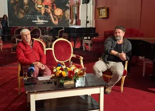 Două persoane stau așezate în fotolii roșii lângă o masă pe care se află un buchet de flori și microfoane într-o încăpere cu podea roșie.