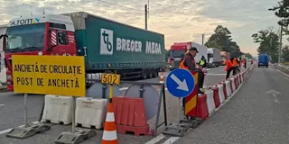 Un semn galben cu text negru stă lângă un camion mare, un con de trafic portocaliu și bariere de plastic alb-roșii pe un drum.