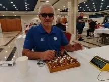 Un bărbat cu ochelari de soare stă la o masă cu un set de șah și un cronometru digital pe fundalul unei săli întinse.