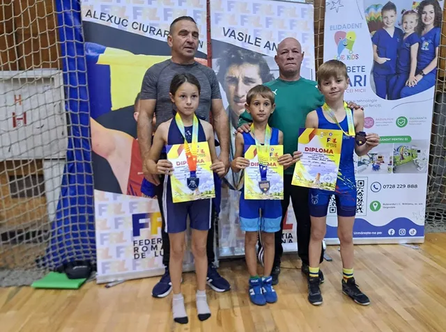 Patru persoane stau în picioare în fața unor panouri publicitare, trei dintre acestea fiind copii care țin diplome și poartă costume de wrestling.