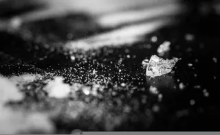 O fotografie alb-negru cu un plan apropiat arată cristale și particule mici împrăștiate pe o suprafață întunecată și lucioasă.