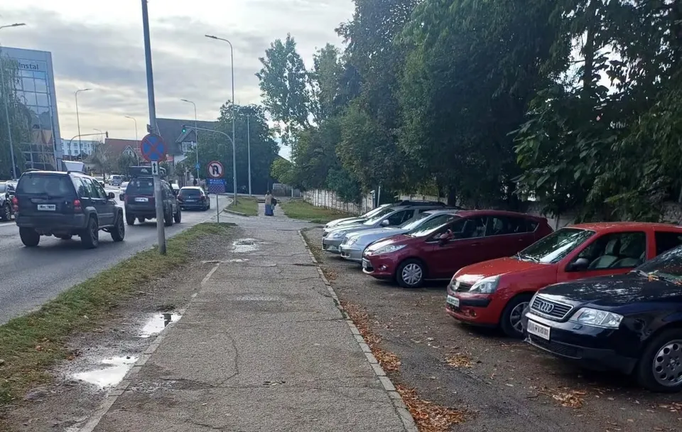 Un trotuar de beton cu fisuri și bălți este situat între o stradă cu mașini circulante și un loc de parcare cu autoturisme în linie.