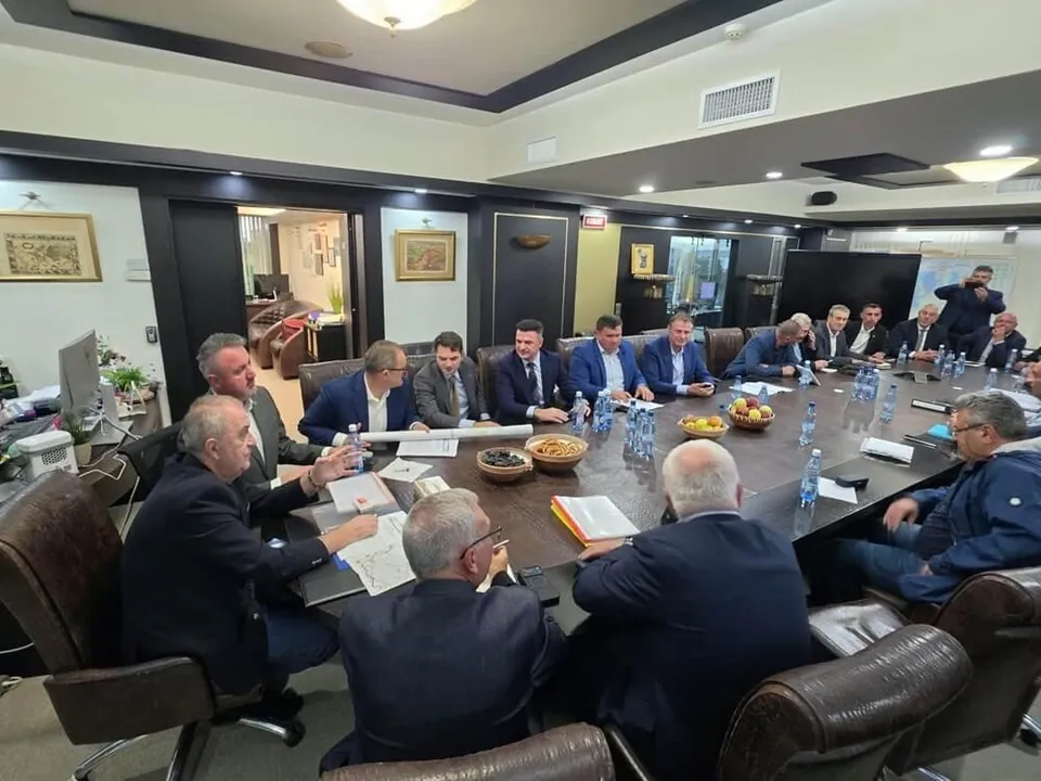 Un grup de bărbați în costume și haine casual stau la o masă lungă de conferință într-o încăpere cu pereți închiși și lumini de tavan.