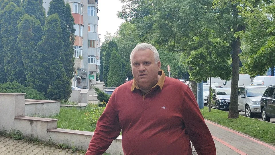 Un bărbat cu părul scurt și deschis la culoare poartă un tricou polo de culoare roșie în fața unor copaci și a unor clădiri.