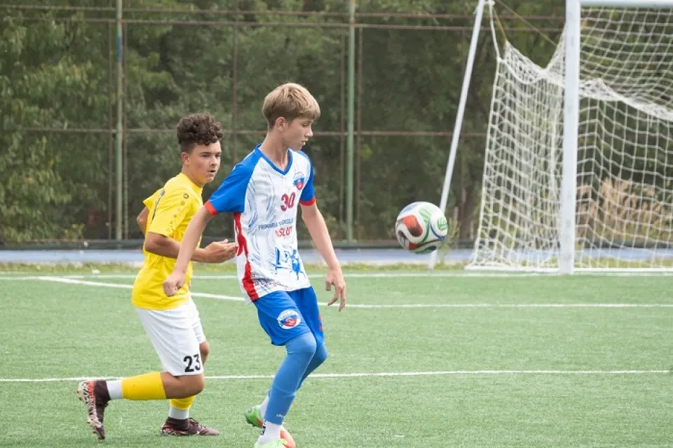 Un jucător într-un echipament albastru și alb urmărește o minge de fotbal pe un teren verde, în timp ce un alt jucător în galben îl urmărește.