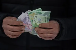 Două mâini țin un teanc de bancnote românești de diferite valori, inclusiv o bancnotă de cinci lei și una de un leu.