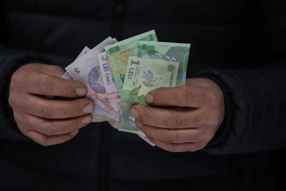 Două mâini țin un teanc de bancnote românești de diferite valori, inclusiv o bancnotă de cinci lei și una de un leu.