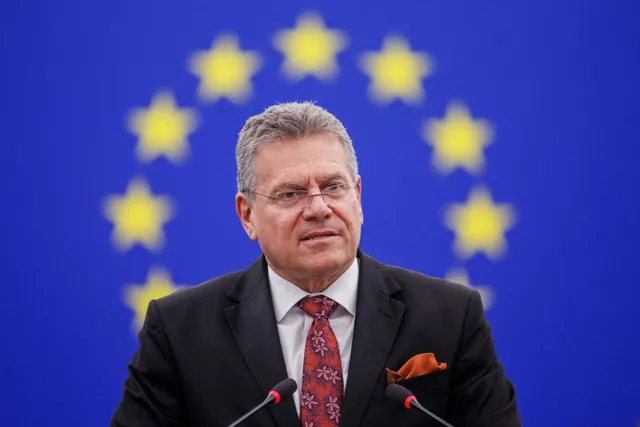 Un bărbat cu ochelari, purtând sacou negru și cravată roșie, stă în fața unui fundal albastru cu stele galbene.