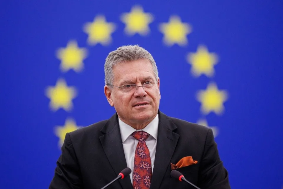 Un bărbat cu ochelari, purtând sacou negru și cravată roșie, stă în fața unui fundal albastru cu stele galbene.