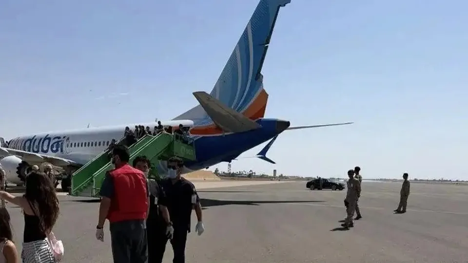 O grupă de persoane coboară pe o scară verde de lângă coada unui avion alb cu dungi albastre și portocalii pe o pistă de aterizare.