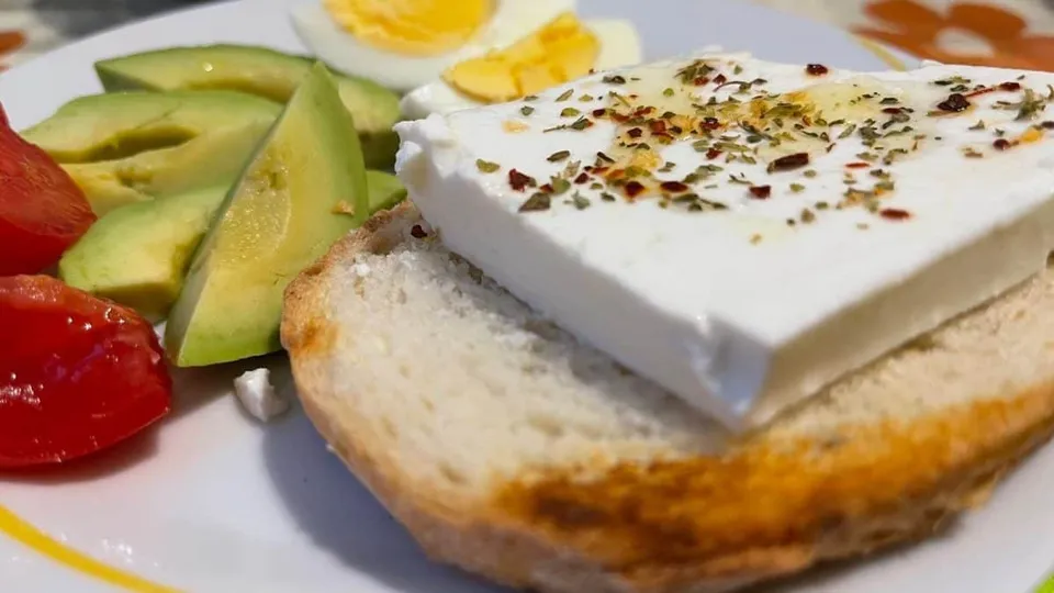 O felie de pâine prăjită este acoperită cu o felie de brânză albă presărată cu condimente, lângă felii de avocado și roșii tăiate.