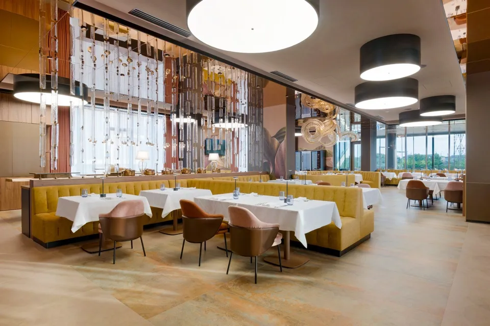 Un restaurant cu mese acoperite cu fețe de masă albe, scaune de culoare roz și canapele galbene sub diverse corpuri de iluminat suspendate.