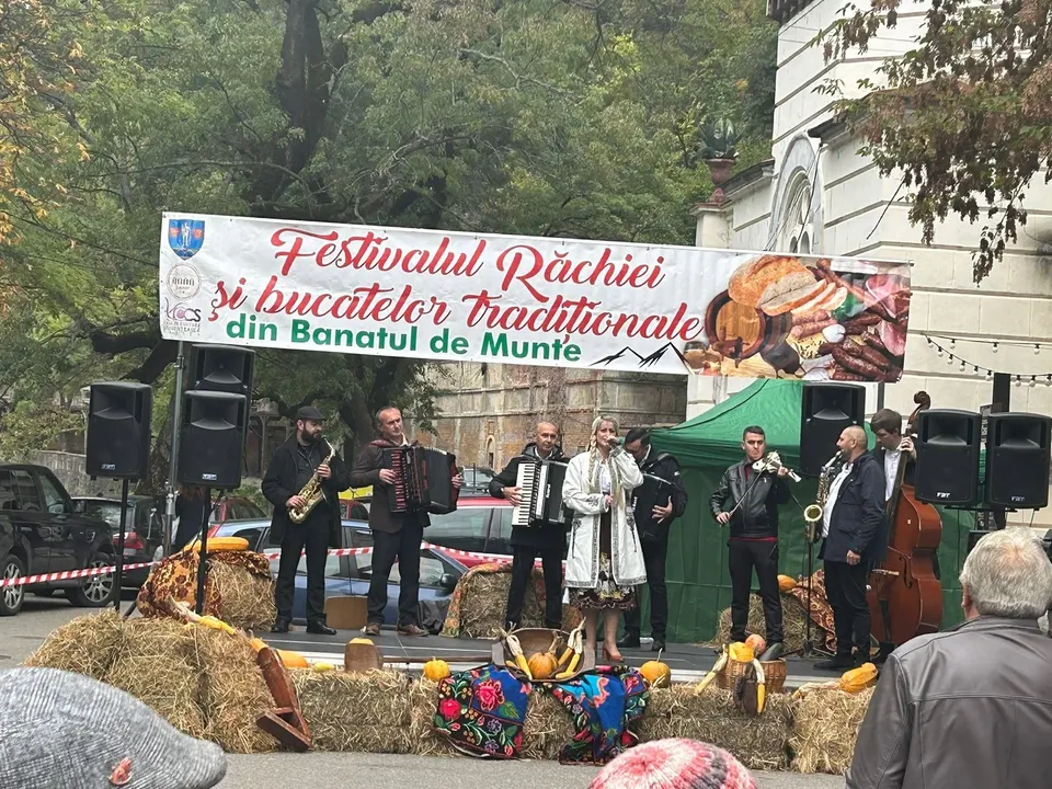 Un grup de muzicieni cântă pe o scenă decorată cu fân, având un banner cu text și imagini de mâncare deasupra lor.