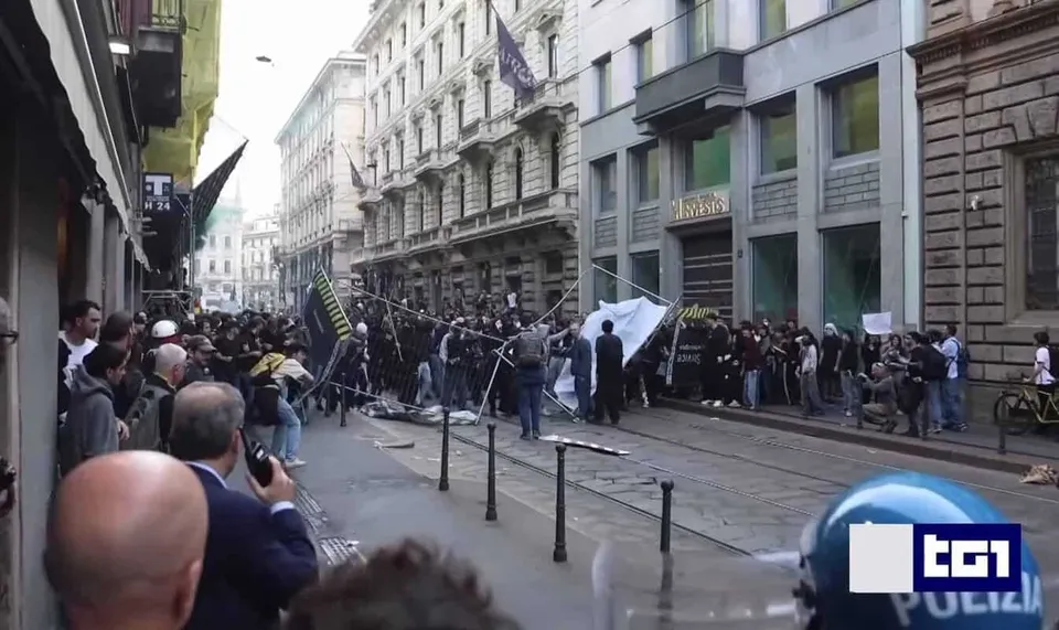 O mulțime de oameni stă pe o stradă îngustă între clădiri înalte, lângă un banner alb și câteva stâlpi metalici pe trotuar.