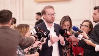 Un bărbat cu ochelari și sacou închis este înconjurat de microfoane și persoane care îi adresează întrebări.