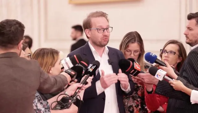 Un bărbat cu ochelari și sacou închis este înconjurat de microfoane și persoane care îi adresează întrebări.