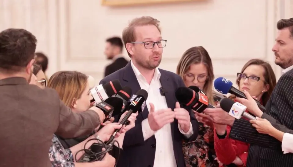 Un bărbat cu ochelari și sacou închis este înconjurat de microfoane și persoane care îi adresează întrebări.