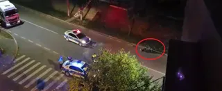 Două mașini de poliție cu luminile aprinse sunt parcate pe o stradă lângă un punct de trecere de pietoni, lângă un grup de persoane.