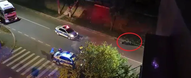 Două mașini de poliție cu luminile aprinse sunt parcate pe o stradă lângă un punct de trecere de pietoni, lângă un grup de persoane.