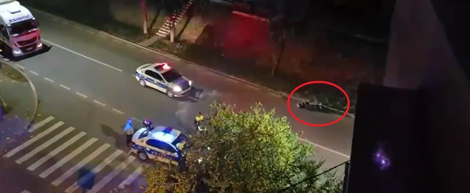 Două mașini de poliție cu luminile aprinse sunt parcate pe o stradă lângă un punct de trecere de pietoni, lângă un grup de persoane.