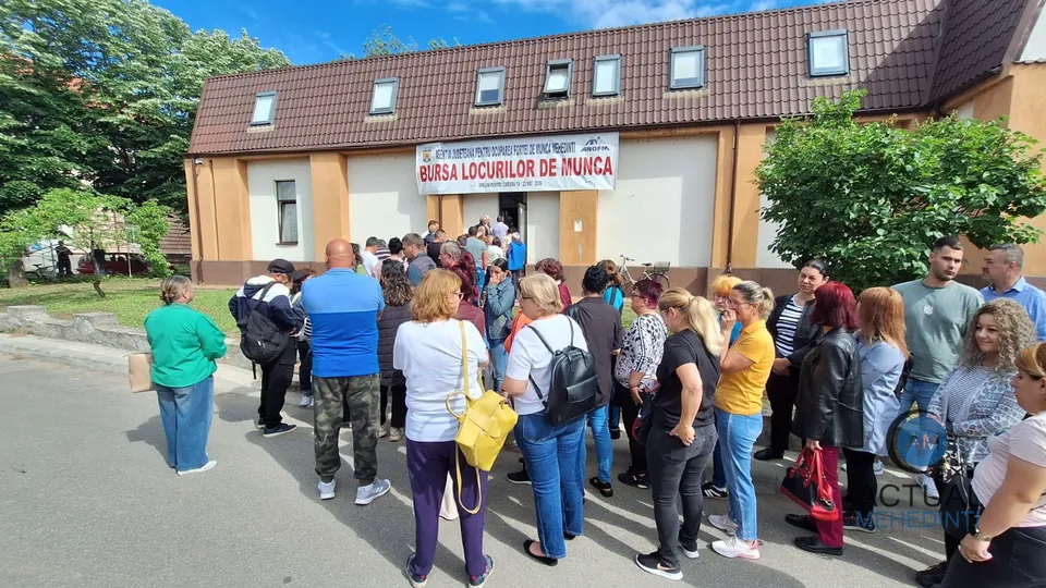 Un grup de persoane stă adunat în fața unei clădiri cu un afiș care menționează bursa locurilor de muncă sub un cer senin.