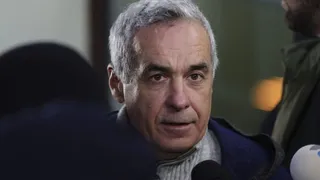 Un bărbat cu păr cărunt poartă pulover și haină albastră, având microfoane în prim-plan și o siluetă neclară în dreapta.