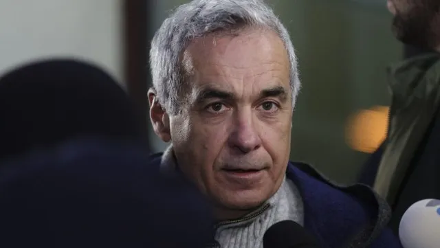 Un bărbat cu păr cărunt poartă pulover și haină albastră, având microfoane în prim-plan și o siluetă neclară în dreapta.