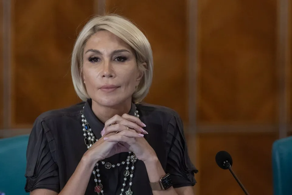 O femeie cu părul blond scurt poartă o cămașă gri închis și un colier cu perle, stând cu mâinile împreunate sub bărbie.