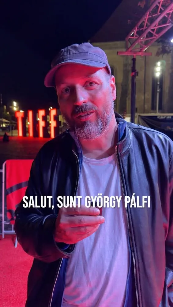 Un bărbat cu barbă, purtând o șapcă, o jachetă neagră și un tricou alb, stă în fața unor litere roșii luminoase pe fundal întunecat.