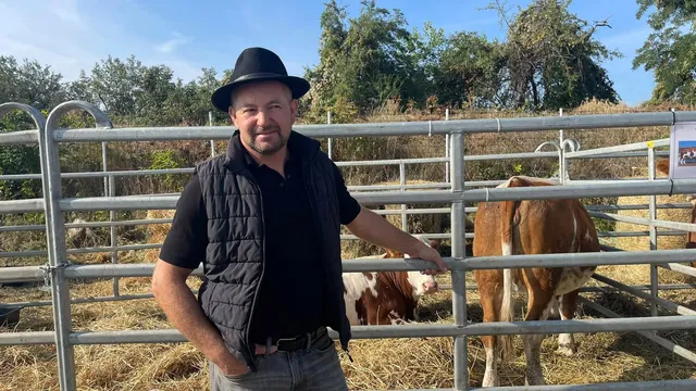 Un bărbat cu pălărie neagră și vestă neagră stă în fața unui gard metalic de fermă lângă niște animale.